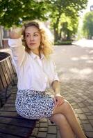 beautiful middle age woman in 70s, 80s style clothes on a bench in a sunlit avenue photo