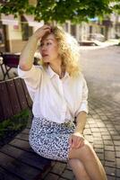 beautiful middle age woman in 70s, 80s style clothes on a bench in a sunlit avenue photo