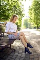 beautiful middle age woman in 70s, 80s style clothes on a bench in a sunlit avenue photo