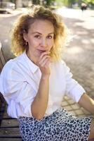 beautiful middle age woman in 70s, 80s style clothes on a bench in a sunlit avenue photo