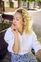 beautiful middle age woman in 70s, 80s style clothes on a bench in a sunlit avenue photo