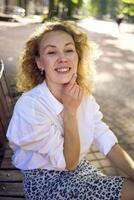 beautiful middle age woman in 70s, 80s style clothes on a bench in a sunlit avenue photo