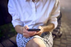 un hermosa medio años mujer en años 70, Años 80 estilo ropa es mirando a su teléfono inteligente en un banco foto