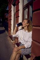 middle age woman in 70s, 80s style clothes with a coffee stain on her shirt drinks coffee sitting on the windowsill photo