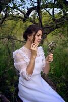 beautiful woman in white vintage dress with train and holding vintage mirror in spring garden at sunset photo