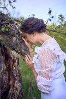 un hermosa mujer en un blanco Clásico vestir con un tren es acariciando un árbol dañado por un tormenta foto