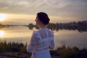un mujer en un blanco Clásico vestir en el apuntalar de un lago a puesta de sol foto