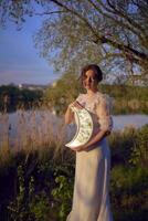 a woman in a white vintage dress on the shore of a lake at sunset holds a moon shaped mirror photo