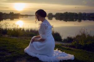 un mujer en un blanco Clásico vestir en el apuntalar de un lago a puesta de sol foto