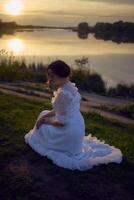 un mujer en un blanco Clásico vestir en el apuntalar de un lago a puesta de sol foto