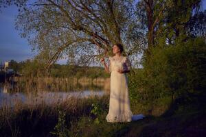 a woman in a white vintage dress on the shore of a lake at sunset photo