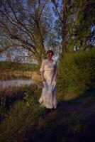 un mujer en un blanco Clásico vestir en el apuntalar de un lago a puesta de sol foto