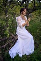 beautiful woman in white vintage dress with train and holding vintage mirror in spring garden at sunset photo