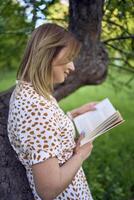 mujer en un pastel vestir lee un libro cerca un árbol en el jardín foto