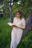 mujer en un pastel vestir lee un libro cerca un árbol en el jardín foto