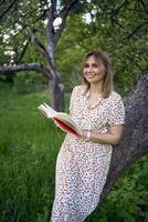 mujer en un pastel vestir lee un libro cerca un árbol en el jardín foto