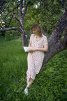 mujer en un pastel vestir lee un libro cerca un árbol en el jardín foto