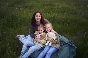 a mother with two children and their toys hug while sitting on a blanket in a field photo