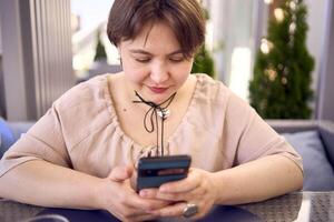 plus size woman in peach fuzz dress chooses a dish in a restaurant via QR code in smartphone, water with lemon and glasses in front of her photo