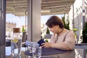 más Talla mujer en melocotón pelusa vestir elige un plato en un restaurante vía qr código en teléfono inteligente, agua con limón y lentes en frente de su foto