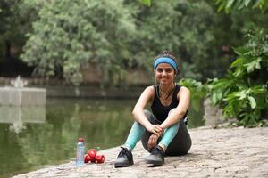 sonriente atractivo dama en ropa de deporte con auriculares sentado en parque foto