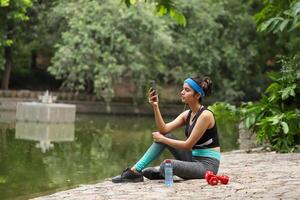 Fit Beautiful Woman Looking At Phone Sitting On Floor With Bottle And Dumbbells photo