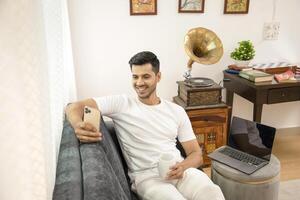 Hot Man Smiling While Looking At His Mobile Phone Sitting In Front Of Laptop On Sofa photo