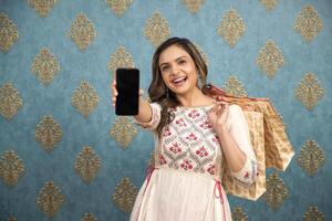 A Cheerful Indian Wear Woman Holding Shopping Bags While Showing The Camera Her Mobile Screen photo