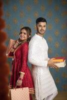 un valores foto de un amor verdadero Pareja posando para el cámara mientras participación diwali regalos en su manos y en pie espalda a espalda en tradicional indio atuendo