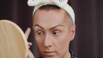 Young male performer in a drag queen makeup, applying eyeshadows with a finger while looking in the hand mirror in the dressing room during preparation for the performance video