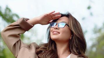 Young asian woman in glasses looking up, shading the eyes with her hand and smiling on the sky background on a sunny day outdoors in the city park close up video