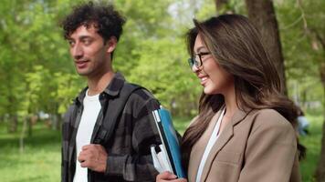 Young smiling asian woman with documents in hands walking on an alley of the city park with a young man in casual clothes and discussing work issues close up video