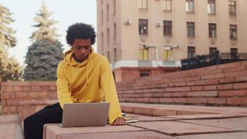 jung konzentriert schwarz Programmierer im beiläufig Kleider Arbeiten auf ein Laptop während Sitzung draußen auf Treppe in der Nähe von das Gebäude. Konzept von flexibel Fernbedienung Arbeit video