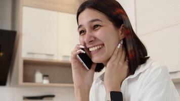 Young attractive woman in casual clothes standing in the cozy kitchen at home, talking with a friend on a smartphone and cheerfully laughing close up video