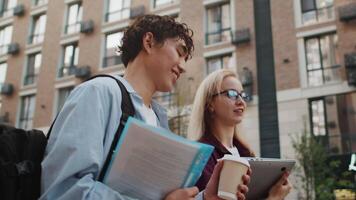 Young smiling caucasian woman with laptop in hands walking on a city street with a young asian man with documents and backpack and discussing work issues close up video