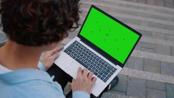 Young asian man with curly hair using a laptop with a mockup green screen when he is sitting on the stairs outdoors in the city street, back view video