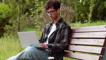 Young man in casual clothes sitting with a laptop on the bench in a city park and working outdoors with a focused face. Concept of flexible remote work video