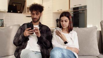 Young couple sitting on the couch at home with their smartphones and don't pay attention to each other. Concept of difficulties in a couple relationship video