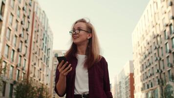 joven atractivo caucásico mujer caminando abajo un calle Entre de muchos pisos edificios en un soleado día cerca arriba, mirando a el teléfono inteligente y sonriente video