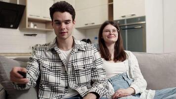 Young man in casual clothes switching channels on TV with a remote control and hugging his smiling girlfriend while sitting on a couch at home. Concept of happiness and harmony in couple relationship video