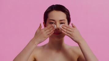 Portrait of a young beautiful woman with clean skin applying nourishing cream on her face with both hands and laughing on a pink background. Concept of skin care for health and beauty video