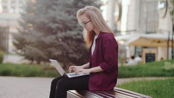 Young attractive caucasian woman in glasses sitting on a bench with a laptop and working with a city building as a backdrop. Concept of flexible remote work video