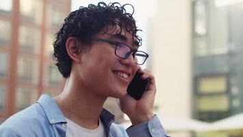 ung asiatisk man med lockigt hår och glasögon stående i de stad gata i solig dag, talande med en vän på en smartphone och glatt leende stänga upp video