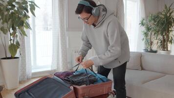 joven hombre en auriculares comprimir arriba un maleta con empaquetado ropa, tendido en un mesa en un acogedor vivo habitación durante preparación a el vacaciones video