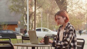 Young caucasian woman in casual clothes drinking coffee from a paper cup and working on a laptop, sitting at the table in the outdoor cafe on a city street video