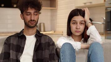 Young upset man sitting on a couch at home next to an offended young woman and looking at the camera after a quarrel. Concept of problems in couple relationship video