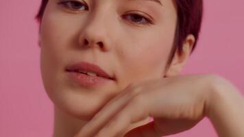 Young attractive woman with clean skin touching her face by the hand close up and coquettishly looking at the camera on a pink background. Concept of skin care for health and beauty video