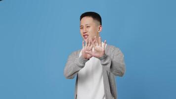 Young asian guy in casual clothes showing disgust, asking to stop by gestures and saying no while standing on a blue background in the studio video