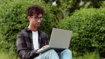 Young man in casual clothes and glasses sitting with a laptop in a city park and working outdoors with a focused face. Concept of flexible remote work video