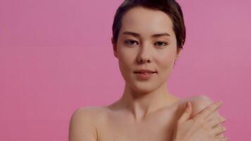 Young beautiful woman with clean skin touching her shoulder and looking at the camera on a pink background close up. Concept of skin care for health and beauty video